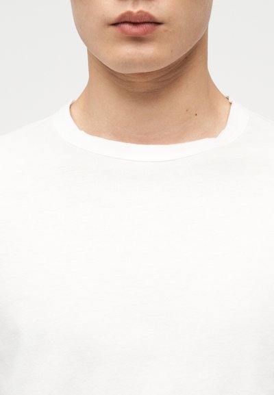 Εγκαρσία λήψη από το κάτω μέρος του προσώπου και τον λαιμό ενός ατόμου που φοράει απλό λευκό μπλουζάκι με στρογγυλή λαιμόκοψη, με φόντο λευκό.