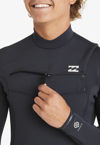 Czarny piankowy skafander z frontowym zamkiem, gładką teksturą i kontrastowym logo marki na klatce piersiowej. Dopasowany krój podkreśla górną część ciała.