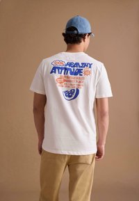 Wit katoenen T-shirt met kleurrijke tekstafbeelding op de achterkant, met blauwe en rode letters, gedragen met beige broek en een blauwe pet.