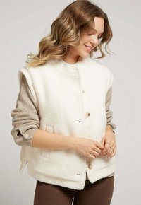 Gilet en polaire blanc avec un col rond, fermeture à boutons sur le devant et poches latérales. Présente un pull beige en dessous, mettant en valeur des manches en tricot texturé.