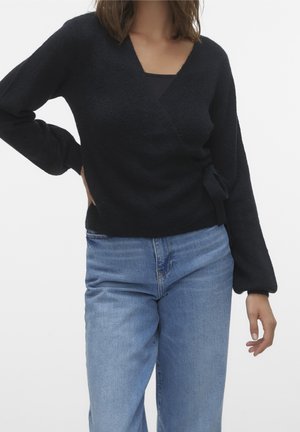 Black wrap sweater made of textured knit fabric. Features a V-neck, long sleeves, and a side tie detail. Paired with light blue jeans.