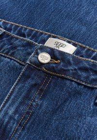 Jeans de mezclilla con un detalle de costura en color cobre, botón metálico plateado y etiqueta de tela con un logotipo negro sobre un fondo blanco.