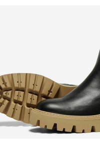 Botas de cuero negras hasta el tobillo con una suela de goma beige gruesa que presenta profundas ranuras y un diseño de punta redondeada.