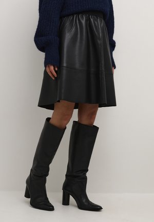 Jupe en cuir noir avec une taille élastique, longueur genou, associée à des bottes noires à talons montant jusqu'aux genoux. Le pull est en tissu texturé de couleur bleu foncé.