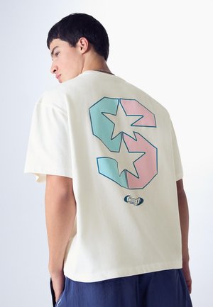 Młody mężczyzna w kremowej koszuli z dużą niebiesko-różową gwiazdą i cyfrą „3” na plecach, w połączeniu z niebieskimi szortami na jednolitym tle.