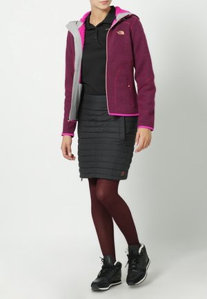 Paarse fleece jas met grijze capuchon, roze voering, zwarte gewatteerde rok, bordeauxrode maillots en zwarte sneakers met bontafwerking.