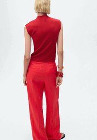 Haut sans manches rouge à col montant associé à un pantalon ample en lin rouge. Accessoires comprenant des bracelets rouges. Texture douce, design minimaliste.