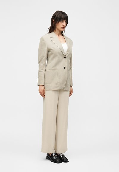 Γυναίκα όρθια που φοράει μπεζ σακάκι με δύο κουμπιά και τσέπες, μπεζ παντελόνες με φαρδύ πόδι, λευκό τοπ και μαύρα loafers.