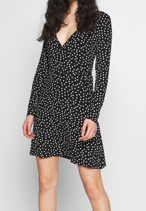 Femme portant une robe noire à manches longues avec des pois blancs, un décolleté en V et une jupe légèrement évasée, debout devant un fond uni.