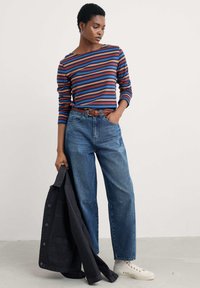 Ženska oblečena v striped dolg rokav top, hlače z visokim pasom, bele teniske, drži črno jakno in stoji ob enobarvni svetli ozadju.