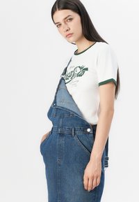 Tumedastest sinistest denimist tööriided reguleeritavate rihmadega, mille all on valge särk, millel on roheline tekst ja äärised. İrge disain taskutega.