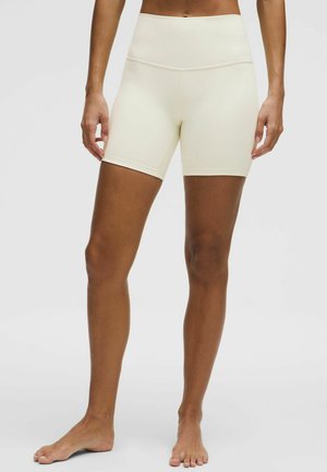 Kvinde model iført højtaljede, cremefarvede biker-shorts, stående barfodet mod en ensartet lys grå baggrund.