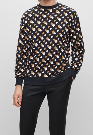 Homme portant un pull noir avec un motif géométrique marron, blanc et noir, associé à un pantalon noir, debout devant un fond uni.