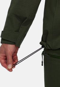 Dettaglio di giacca verde che mostra una mano che tira un cordino nero in vita, con una tasca con zip visibile sul capo inferiore.
