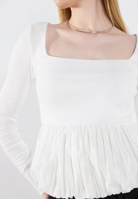 Bijela bluzica dugih rukava s kvadratnim izrezom i nabranim, teksturiranim peplum dizajnom. Glatka, mekana tkanina kontrastira s volanastim dnom.