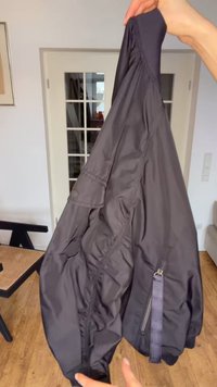 Des mains tenant une veste légère noire à l'intérieur, montrant une manche avec une poche zippée et une poche à rabat boutonnée à proximité.