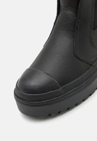 Botín de cuero negro con una puntera redonda, parte superior texturizada y suela gruesa con patrones de tracción. El botín tiene detalles de costura visibles.