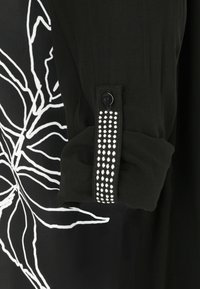 Schwarzes Oberteil mit einem weißen abstrakten Blattmuster, versehen mit einem Ärmelabschluss, der mit einer Reihe dekorativer klarer Perlen und einem passenden Knopf geschmückt ist.