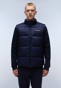 Marinblå pufferväst med hög krage, dragkedjestängning och logotyp. Slät textur, väldefinierad quiltning, kombinerad med en mörk topp och byxor.