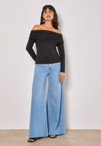 Schwarzes Carmen-Oberteil mit langen Ärmeln, kombiniert mit weit geschnittenen, hellblauen Jeans. Glatte Stoffstruktur, minimalistisches Design und keine sichtbaren Muster.