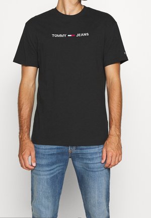 T-shirt en coton noir avec un logo blanc "TOMMY" et des accents rouges et blancs entre "TOMMY" et "JEANS". Manches courtes, col rond.