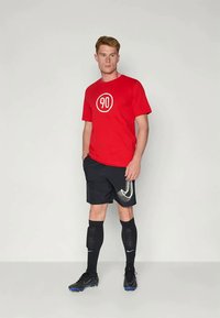 Mandlig atlet står iført en rød "90" T-shirt, sorte shorts med stor swoosh, sorte knæstrømper og sorte støvler med blå såler.