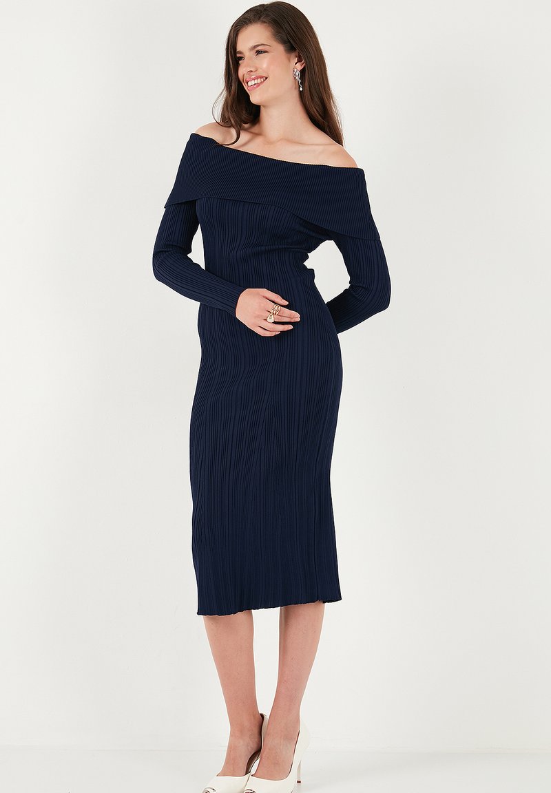Abito midi a coste blu navy con scollatura off-the-shoulder e maniche lunghe. Texture liscia, design aderente e orlo dritto. Paio di tacchi bianchi.