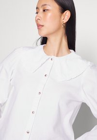 Camisa branca de botão com gola franzida e mangas longas, com botões rosa e um tecido leve e suave.