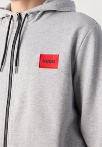 Sweat à capuche gris clair zippé avec cordons noirs et un patch carré rouge portant l'inscription "HUGO" sur la poitrine.