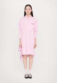 Ροζ καρό φόρεμα με κολάρο, κλείσιμο με κουμπιά και τσέπη στο στήθος που διαθέτει το λογότυπο "Fiorucci". Μοντέλο που κρατάει ένα ταιριαστό ροζ αξεσουάρ.