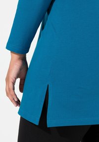 Maglietta turchese a maniche lunghe con uno spacco laterale all'orlo e texture liscia, abbinata a leggings neri.