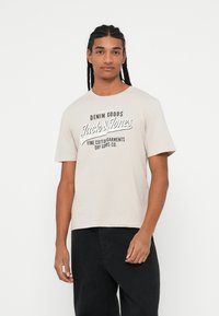 Béžové bavlněné tričko s velkým černým grafickým potiskem s nápisem "Jack & Jones" a kulatým výstřihem. Nosené s černými kalhotami.