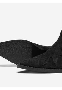 Botte en daim noir avec un bout pointu, surface texturée, un accent de design ondulé subtil et une semelle en caoutchouc plate et mouchetée.