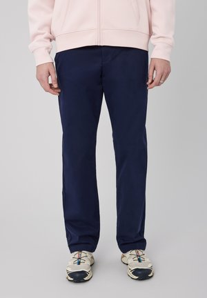 Homme portant un pantalon bleu marine, des baskets beiges et noires, et un sweat-shirt rose clair zippé, debout devant un fond uni.