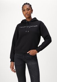 Sweat à capuche noir court avec le logo blanc "TOMMY HILFIGER". Dispose de cordons de serrage et de poignets côtelés. Assorti à un jean foncé.