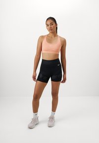 Top sportivo color pesca chiaro con spalline sottili e logo bianco, abbinato a pantaloni corti neri a vita alta con "Nike Pro" in grigio. Indossato con sneakers rosa chiaro.