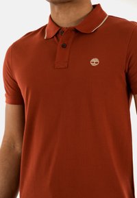 Polo in arancione ruggine, realizzato in tessuto testurizzato. Presenta una rifinitura beige sul colletto, due bottoni neri e un logo discreto sul petto.