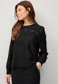 Haut en dentelle noire à manches longues, présentant un motif de formes diamantées et d'encoches, col rond et coupe décontractée.