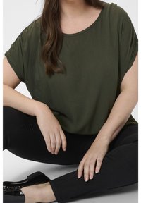 Femme aux longs cheveux bruns assise en tailleur, portant un haut vert olive, un pantalon noir et des mocassins noirs brillants.