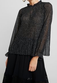 Blouse noire transparente à pois blancs, encolure à volants, manches longues et poignets élastiques. Associée à une jupe noire unie avec bordure en dentelle.