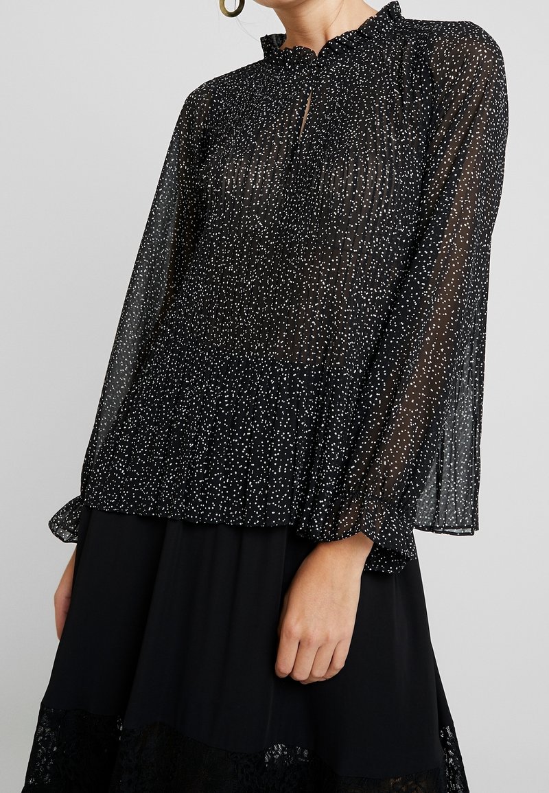 Blouse noire transparente à pois blancs, encolure à volants, manches longues et poignets élastiques. Associée à une jupe noire unie avec bordure en dentelle.
