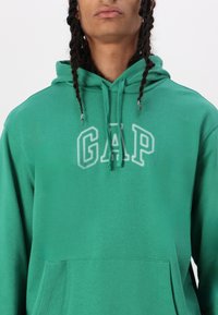 Žalias medvilnės džemperis su priekine kišene ir baltu "GAP" logotipu ant krūtinės. Su raišteliu reguliuojama gobtuvą ir švelni tekstūra. Minimalistinis dizainas.