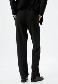 Pantalon noir avec une taille élastique, présentant une texture lisse et un design à jambes droites, associé à des chaussures noires brillantes.