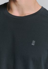 Donkergrijze T-shirt met korte mouwen van katoen. Heeft een ronde hals en een klein wit logo met horizontale lijnen aan de linkerkant.