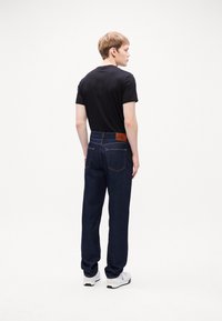 Jeans de denim azul oscuro con un corte relajado, que cuentan con bolsillos traseros y un parche de cuero marrón con el logo en la cinturilla. Llevados con una camiseta negra.