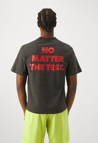 Tmavě šedé bavlněné tričko s výrazným červeným nápisem na zádech "NO MATTER THE TEST." Krátké rukávy a volný střih.