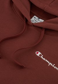 Hoodie marron en coton, avec une poche à l'avant, des cordons de serrage et un logo Champion brodé en blanc. Texture douce.