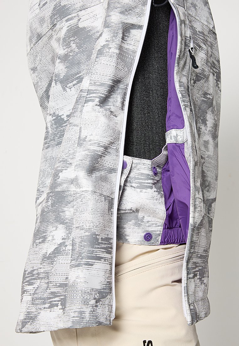 Chaqueta de patrón gris y blanco con forro interior morado, parcialmente desabrochada para mostrar los detalles de broche y cintura elástica sobre pantalones beige.