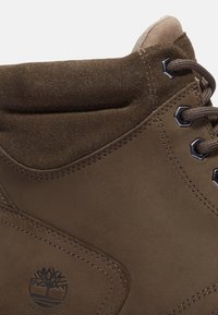Hnědá semišová kotníčková bota s šestihrannými očky, zesíleným prošíváním a voděodolným logem vyraženým na boku. Měkký, texturovaný povrch.