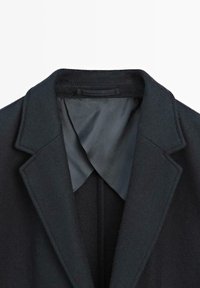 Czarny wełniany płaszcz z wyciętym kołnierzem, z gładką szarym podszewką i wyraźnie zarysowanymi ramionami. Teksturowana powierzchnia z dopasowanymi detalami projektowymi.